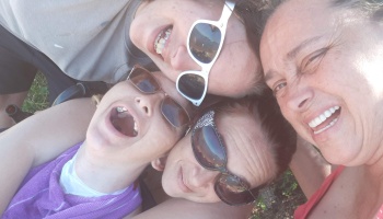 Four happy women wearing sunglasses lean their heads together for a fun, close-up overhead group selfie.