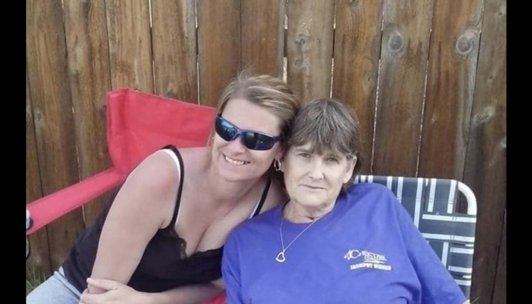 Two women smiling and sitting close together in lawn chairs in front of a wooden fence.