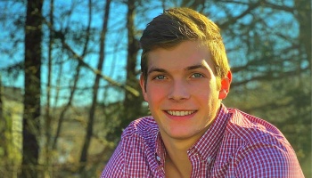 A smiling young man with light brown hair wearing a red checkered shirt outdoors.