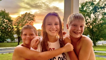 Three smiling children hugging tightly on a porch with a warm sunset glowing in the background.
