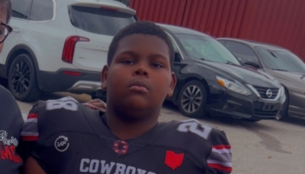 A young boy in a black Cowboys football jersey looking serious outdoors in a parking lot.