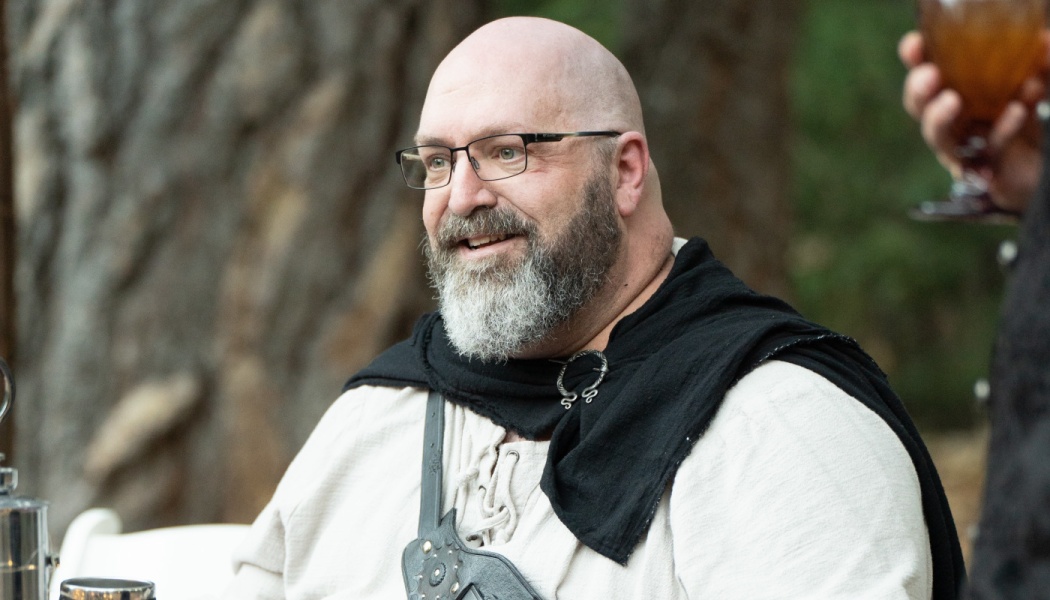 A smiling bald man with a beard and glasses, dressed in a medieval-style tunic and black cloak.