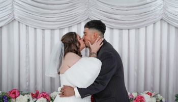 A bride in a veil and her groom in a dark suit kiss passionately against a white draped backdrop with flowers.