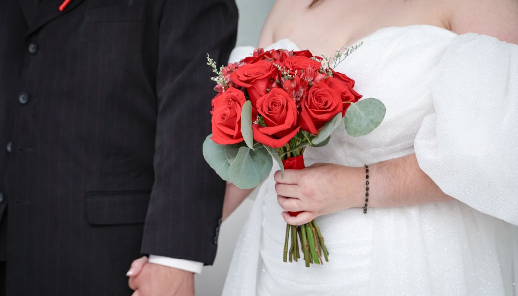 A bride in a white dress holds a vibrant bouquet of red roses while standing next to a groom in a black suit.
