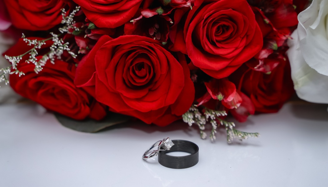 A diamond engagement ring and black wedding band resting in front of a bouquet of deep red roses.
