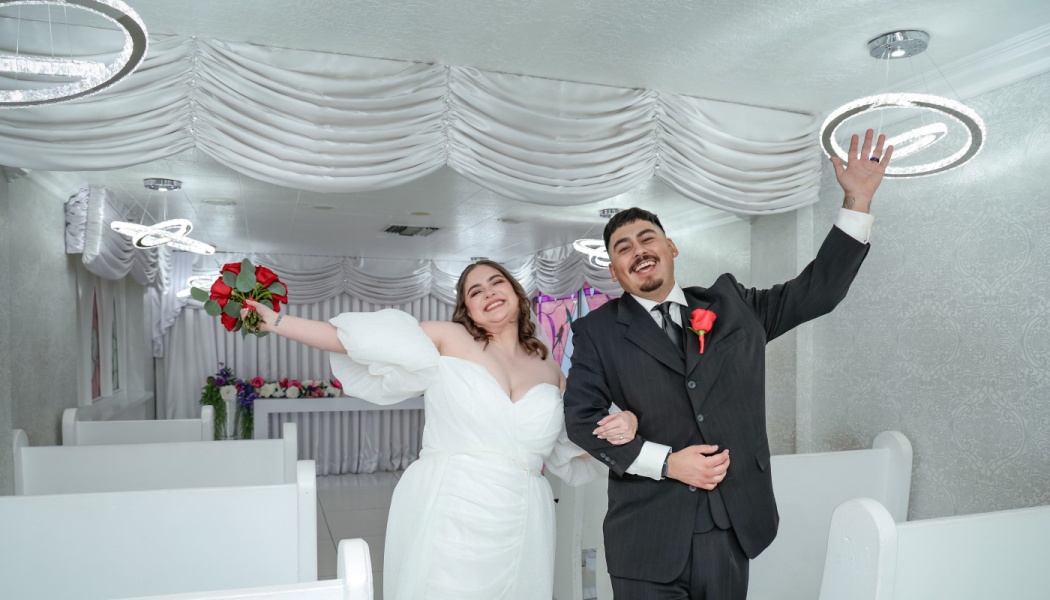 Smiling bride holding red roses and groom waving cheerfully inside a decorated white wedding chapel.