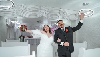 Smiling bride holding red roses and groom waving cheerfully inside a decorated white wedding chapel.