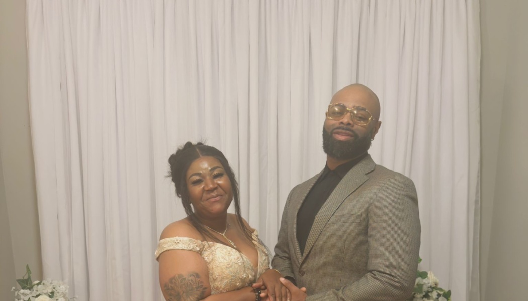 A smiling Black couple in formal attire holding hands in front of a white curtain backdrop.