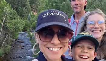 A smiling family of four takes a selfie outdoors with a flowing stream and pine trees in the background.
