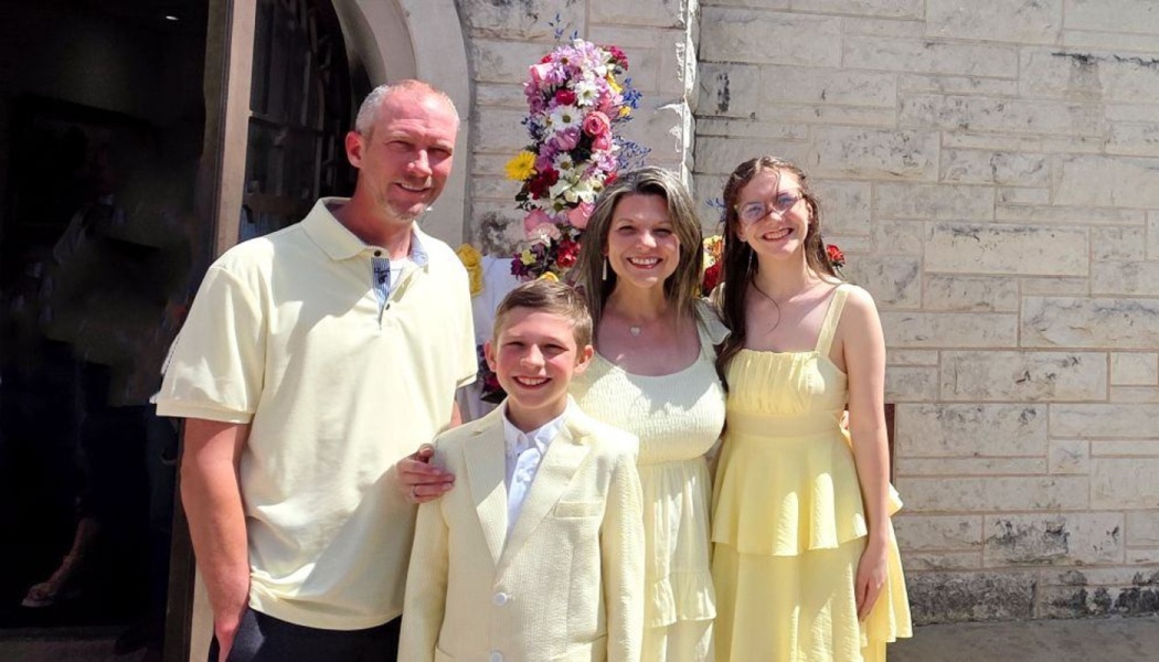 A smiling family of four wearing matching light yellow outfits outdoors in front of a stone building.