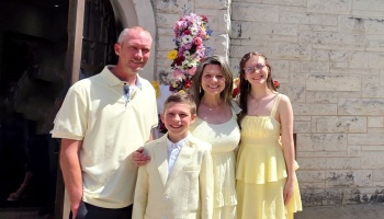 A smiling family of four wearing matching light yellow outfits outdoors in front of a stone building.