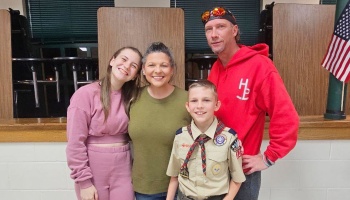 A smiling family of four standing together, featuring a young boy in a Boy Scout uniform.