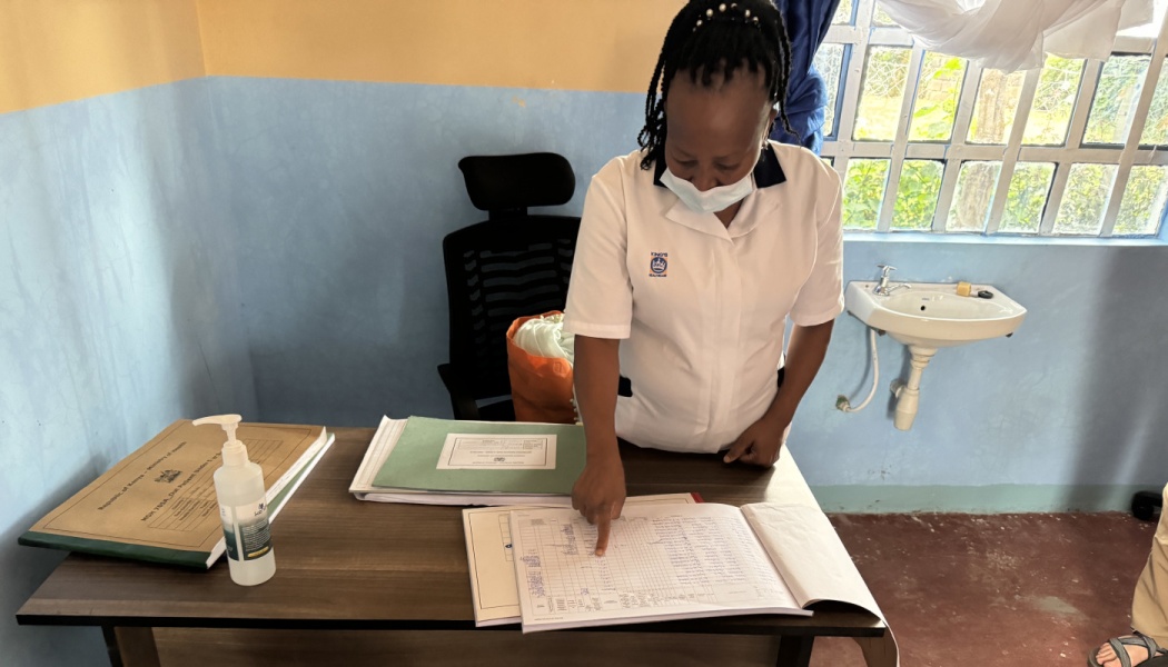 A healthcare worker wearing a mask points to an open medical logbook on a clinic desk.
