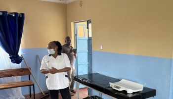 A healthcare worker wearing a face mask stands in a clinic room with an examination table and baby scale.
