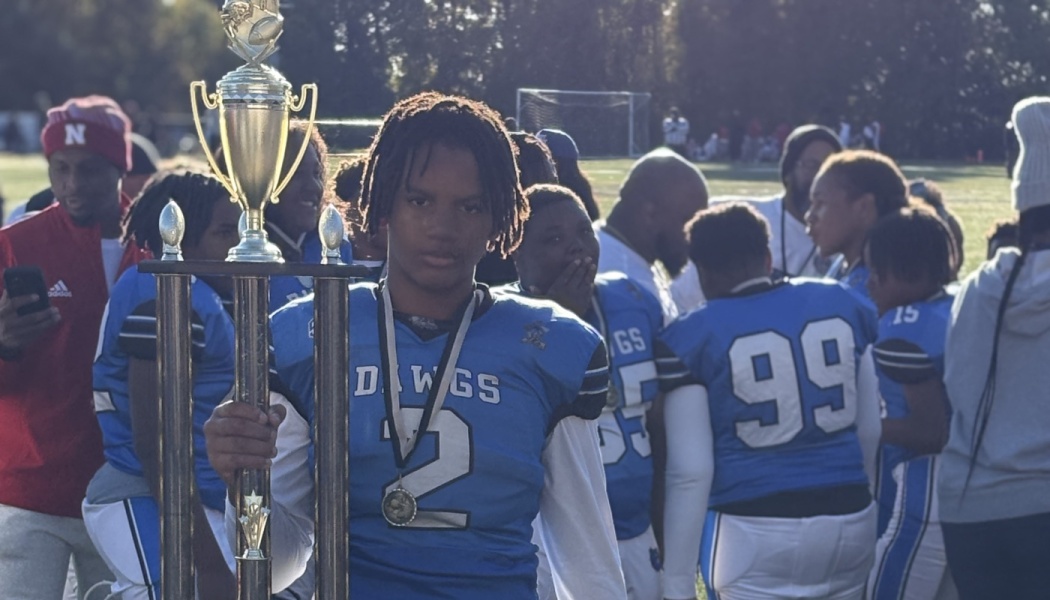 A young football player in a blue jersey holds a tall trophy with a medal around his neck alongside his team.