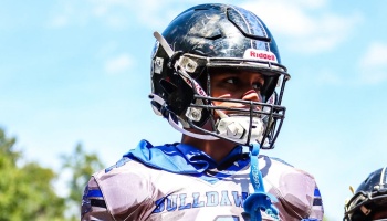 Young football player wearing a black helmet and gray Bulldawgs jersey looking focused under a bright sky.