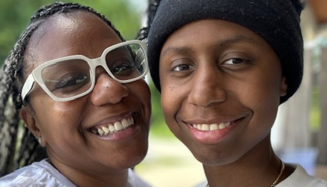 A smiling Black woman in white glasses and a person in a black beanie pose closely together.