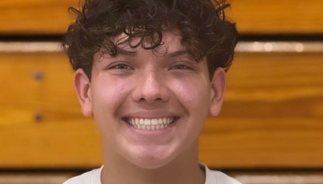A smiling young man with dark curly hair posing in front of wooden gymnasium bleachers.