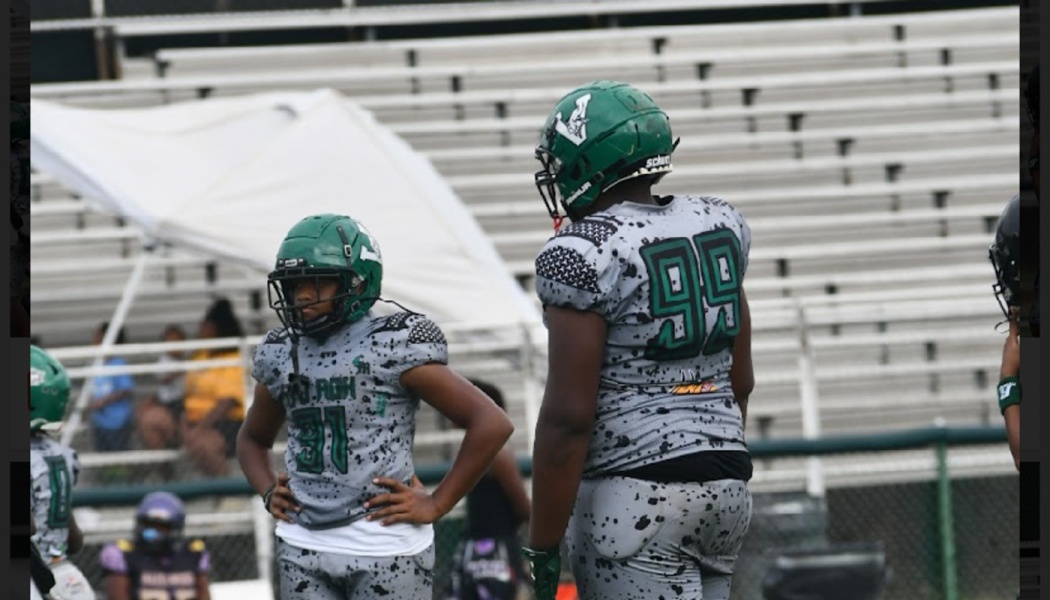 Two young football players in green helmets and grey speckled uniforms stand on the field in front of bleachers.