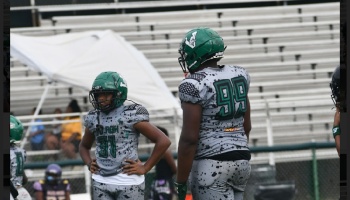 Two young football players in green helmets and grey speckled uniforms stand on the field in front of bleachers.