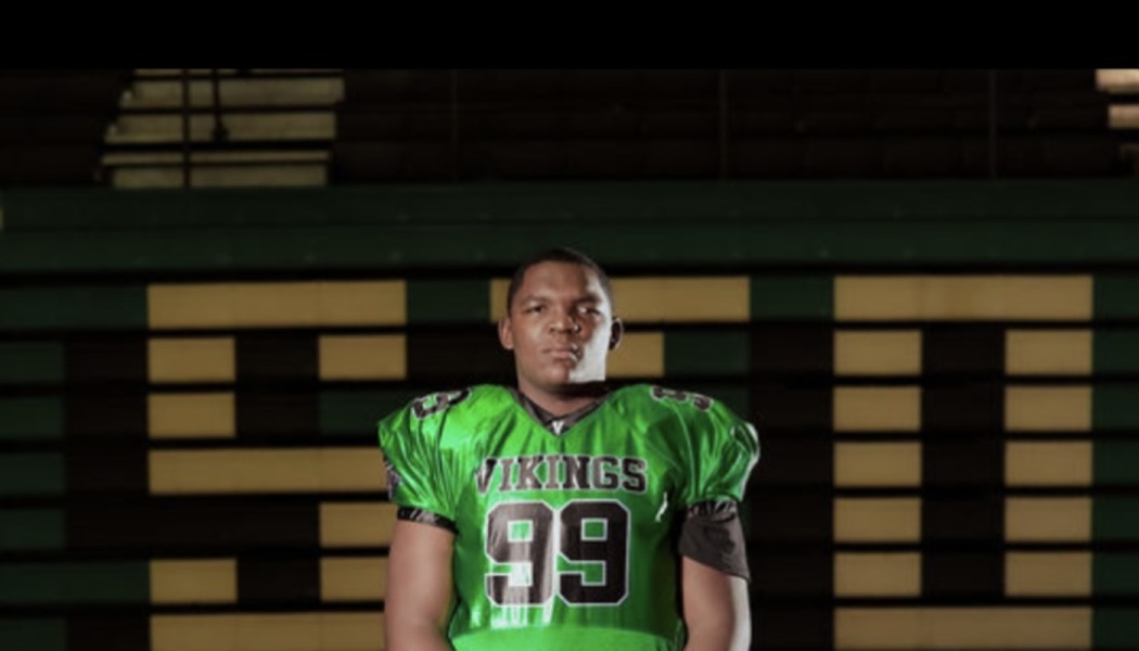 Football player wearing a green Vikings number 99 jersey standing in front of stadium bleachers.