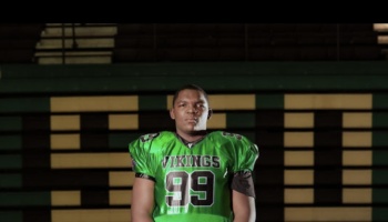 Football player wearing a green Vikings number 99 jersey standing in front of stadium bleachers.