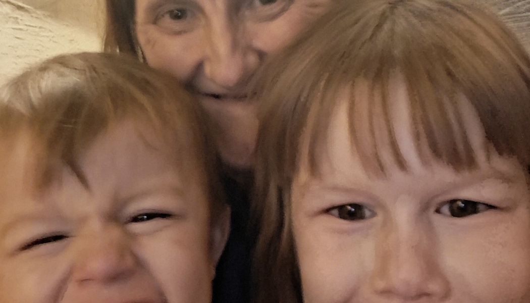 A close-up selfie of a woman smiling behind two young children making faces at the camera.