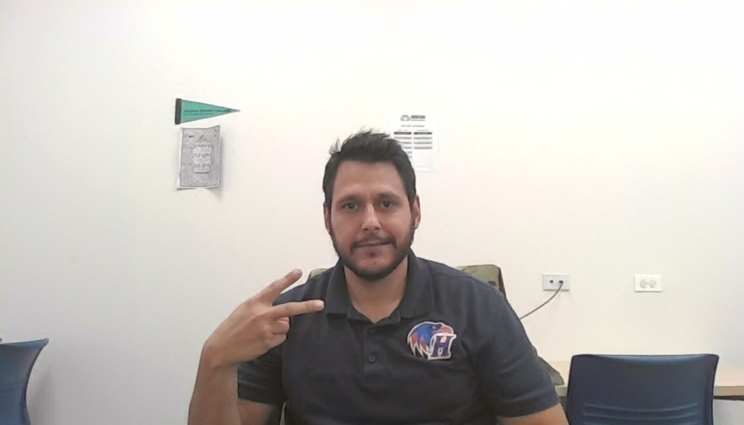 A man with a short beard wearing a dark polo shirt, making a peace sign gesture in an office setting.
