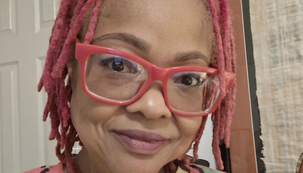Close-up of a smiling Black woman with bright pink dreadlocks and red eyeglasses.