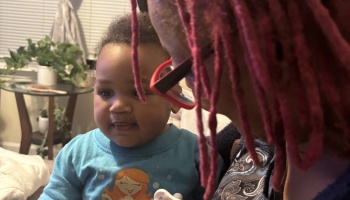 A woman with red locs and glasses looks tenderly at a smiling toddler in a bright blue shirt.