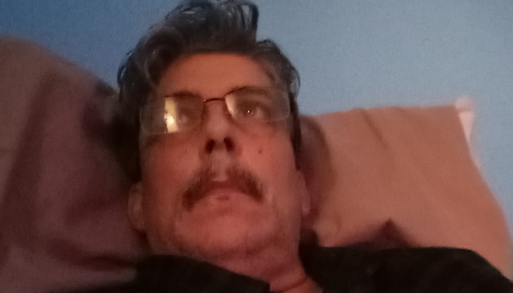 Man with glasses, dark wavy hair, and a mustache resting his head on a pillow.