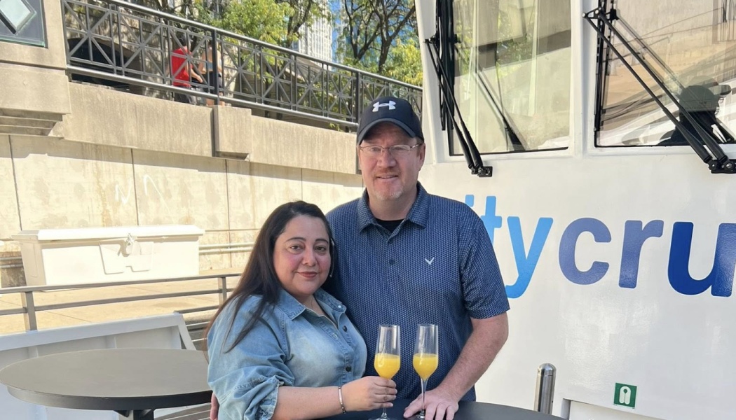 A smiling man and woman pose together on a boat deck, holding champagne flutes with mimosas.