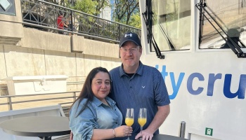 A smiling man and woman pose together on a boat deck, holding champagne flutes with mimosas.