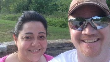 A smiling dark-haired woman and a man wearing a cap and sunglasses taking a selfie outdoors in a park.