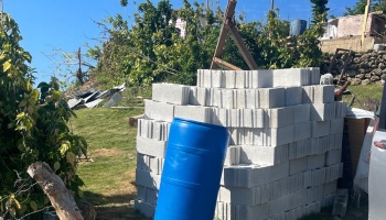 A stack of concrete cinder blocks and a blue plastic barrel outdoors on a grassy hill, ready for rebuilding.