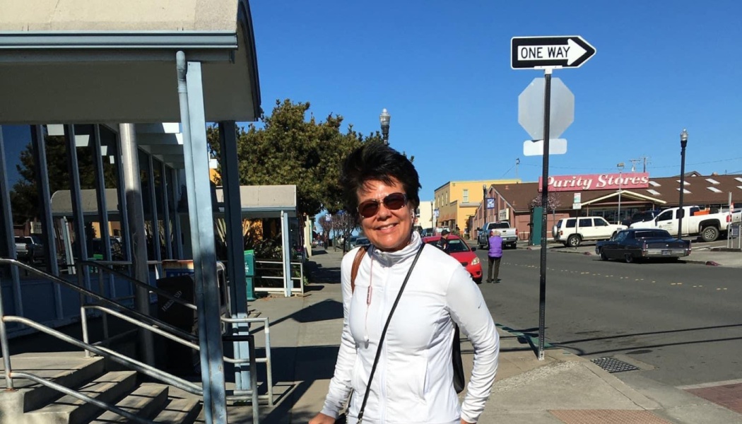 A smiling woman with short dark hair and sunglasses, wearing a white jacket on a sunny town street.