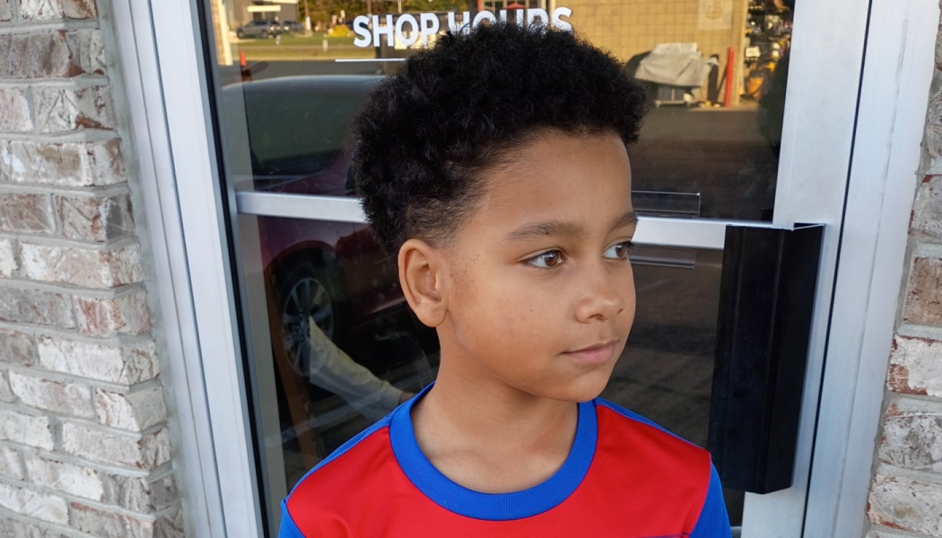 A young boy with short curly hair wearing a red and blue shirt, looking thoughtfully off to the side.