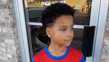 A young boy with short curly hair wearing a red and blue shirt, looking thoughtfully off to the side.