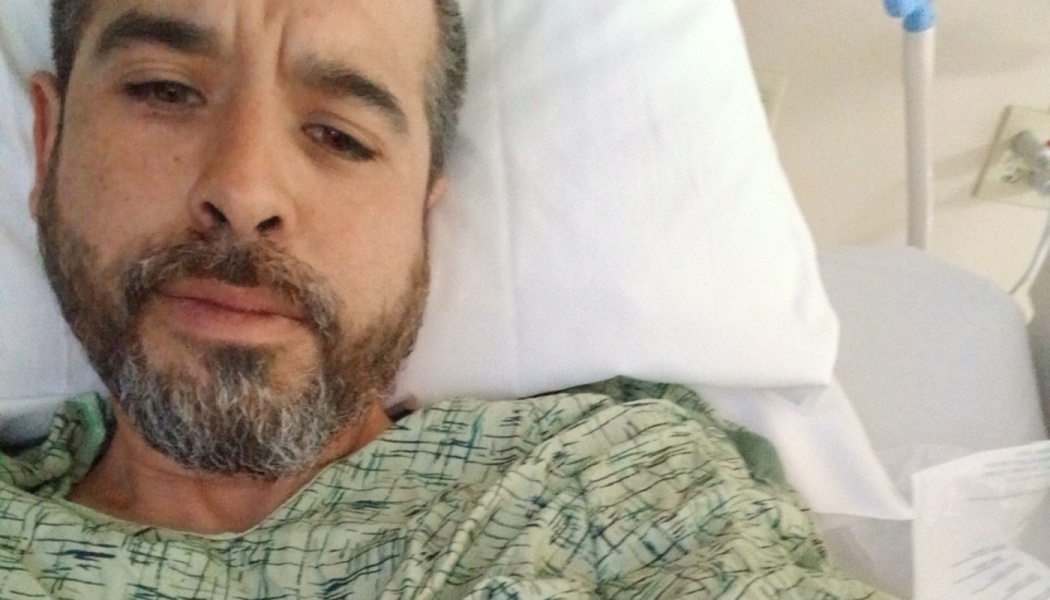 A bearded man wearing a patterned hospital gown rests his head on a white pillow in a hospital bed.
