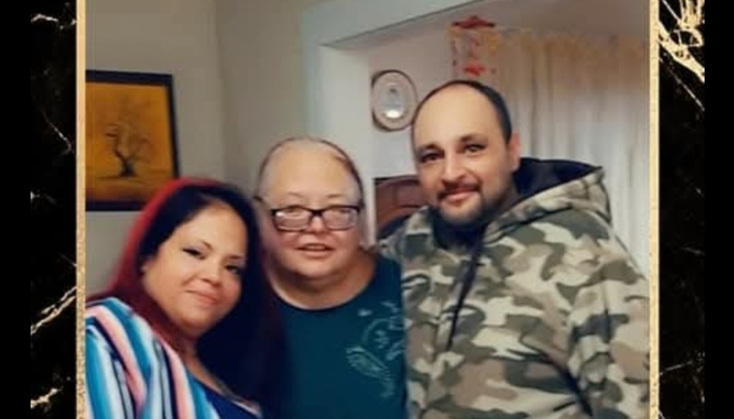 Three smiling adults—two women and a man in a camouflage hoodie—standing close together indoors.
