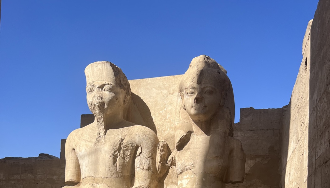 Two ancient Egyptian stone statues of a pharaoh and queen against a clear blue sky.