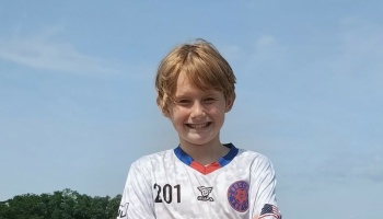A smiling young boy with blonde hair wearing a white youth soccer jersey outdoors against a clear blue sky.