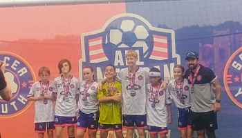 Youth soccer players wearing medals pose with their coach in front of an All-American soccer backdrop.