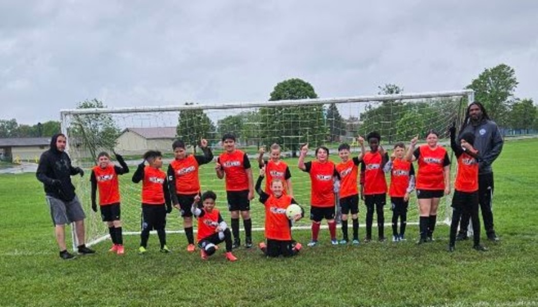 Youth soccer team wearing bright red jerseys poses with coaches in front of a soccer goal on a field.