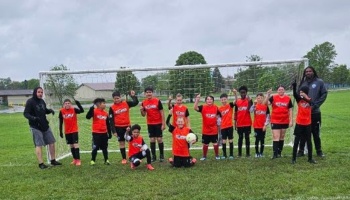 Youth soccer team wearing bright red jerseys poses with coaches in front of a soccer goal on a field.