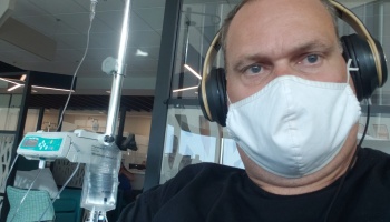 Man wearing a white face mask and over-ear headphones sitting next to an IV drip machine.