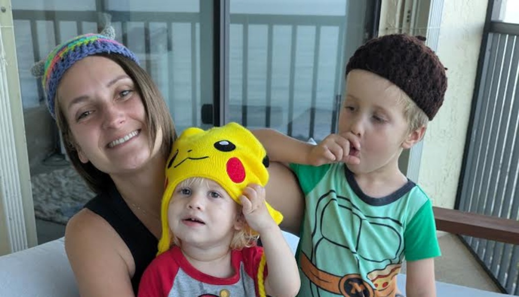 A smiling woman and her two young children wear fun hats, including a yellow Pikachu beanie, on a balcony.