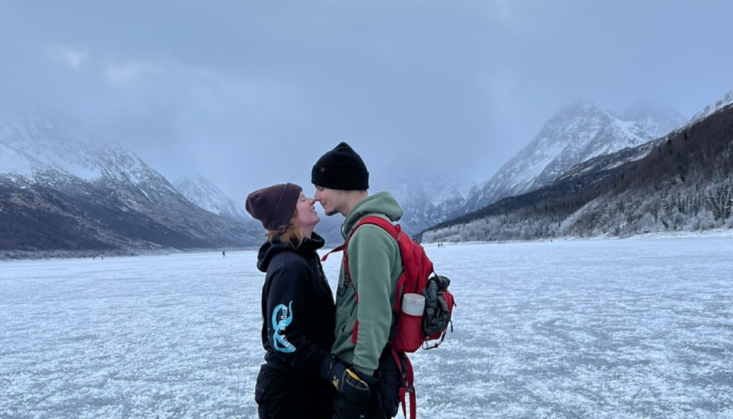 A couple in winter gear touches noses on a frozen lake surrounded by snowy mountains.