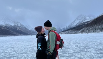 A couple in winter gear touches noses on a frozen lake surrounded by snowy mountains.