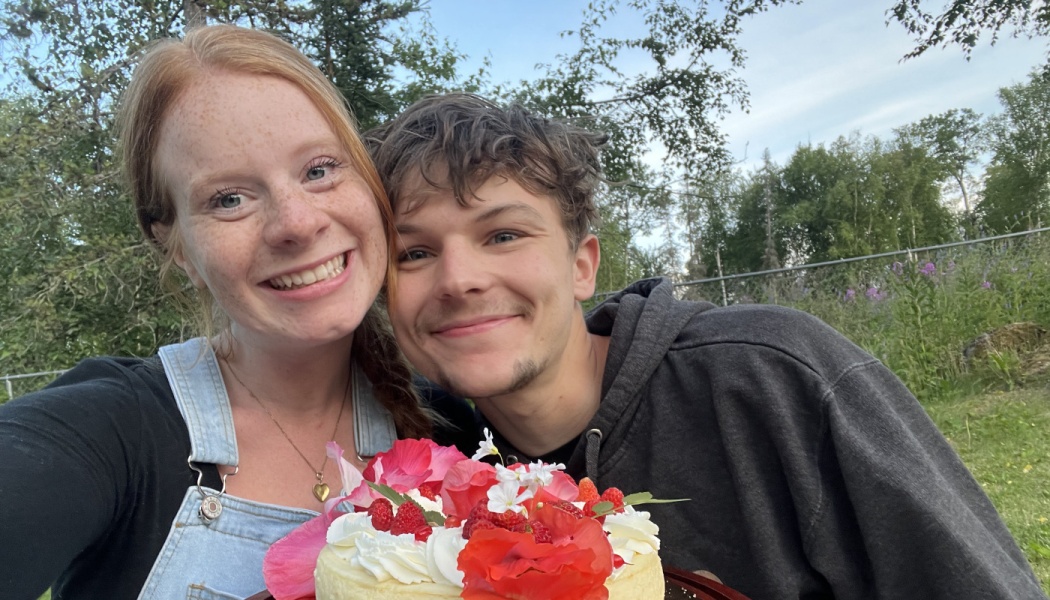 A smiling young couple poses outdoors behind a cake decorated with whipped cream, strawberries, and bright flowers.